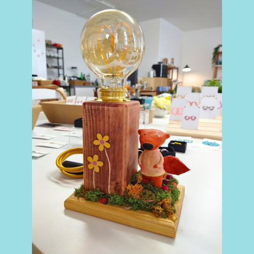 Handgefertigte Tischlampe mit Vintage-Glühbirne in Herzform, Holzsockel, Moosdekoration und Miniaturfigur eines Fuchses – kunstvoll und nachhaltig gestaltet von einem Künstler mit medizinischem Hintergrund. Die Lampe steht auf einem Tisch in einem liebevoll eingerichteten Laden für Kunsthandwerk.