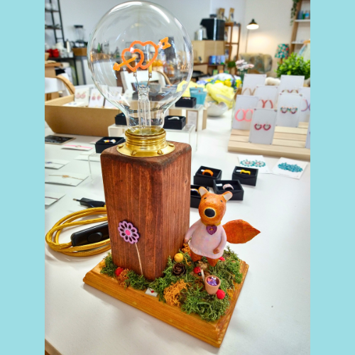 Handgefertigte Tischlampe mit großer Glühbirne in Herzform, auf einem Holzsockel mit Mooslandschaft, Miniaturfuchs in rosa Kleid und liebevollen Details wie Blüten und Pilzen – nachhaltiges Kunsthandwerk aus natürlichen Materialien, fotografiert in einem kreativen Atelier oder Laden.